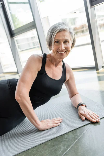 Положительная старшая спортсменка улыбается во время тренировки на фитнес-коврик в тренажерном зале — стоковое фото