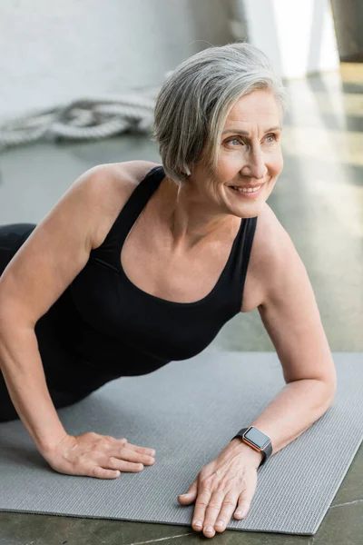 Joyful senior sportswoman smiling while exercising on fitness mat in gym — Stock Photo