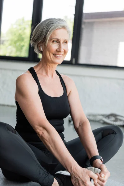 Overjoyed senior sportswoman smiling while stretching on fitness mat in gym — Stock Photo