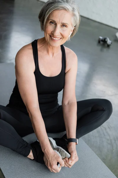 Joyful senior sportswoman smiling while stretching on fitness mat in gym — Stock Photo