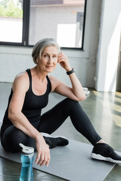 Mujer mayor en ropa deportiva escuchar música y sentarse en la alfombra de fitness en el gimnasio - foto de stock