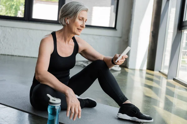 Старшая женщина в спортивной одежде с помощью мобильного телефона и сидя на фитнес-коврик в тренажерном зале — стоковое фото