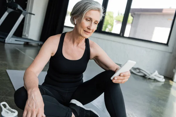 Старшая спортсменка с помощью мобильного телефона и сидя на фитнес-коврик в тренажерном зале — стоковое фото