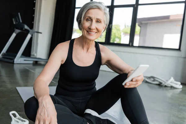 Cheerful sportswoman using mobile phone and sitting on fitness mat in gym — Stock Photo