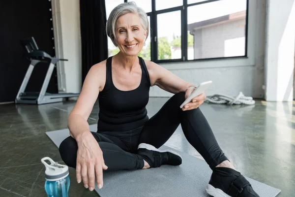 Cheerful sportswoman with grey hair using mobile phone and sitting on fitness mat in gym — Stock Photo