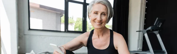 Cheerful sportswoman with grey hair using mobile phone after working out in gym, banner — Stock Photo