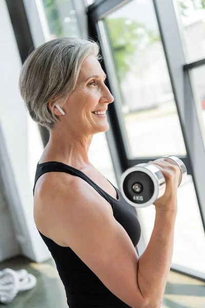 Side view of cheerful senior sportswoman in wireless earphone exercising with dumbbell in gym — Stock Photo