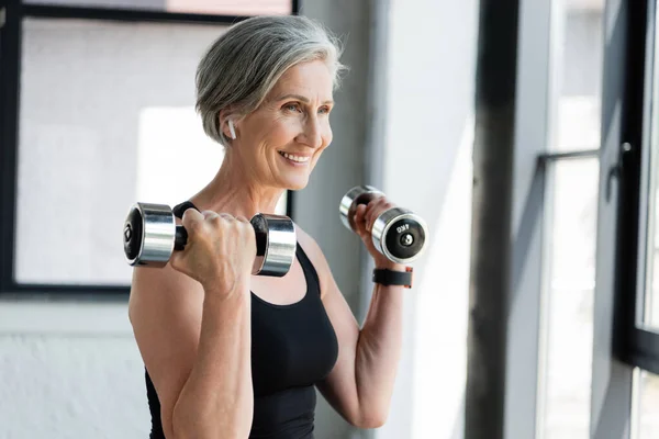 Mujer feliz y mayor en el ejercicio auricular inalámbrico con pesas en el gimnasio - foto de stock