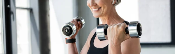 Cropped view of happy retired woman in wireless earphone exercising with dumbbells in gym, banner — Stock Photo