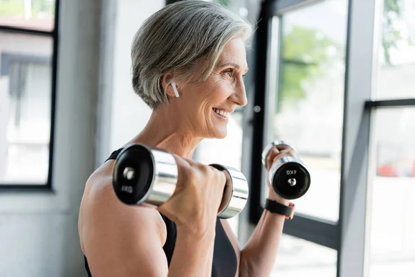Deportista senior feliz en auriculares inalámbricos haciendo ejercicio con pesas en el gimnasio - foto de stock