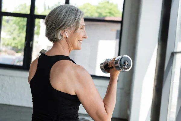 Überglückliche Seniorin mit drahtlosem Kopfhörer trainiert mit Hanteln im Fitnessstudio — Stockfoto