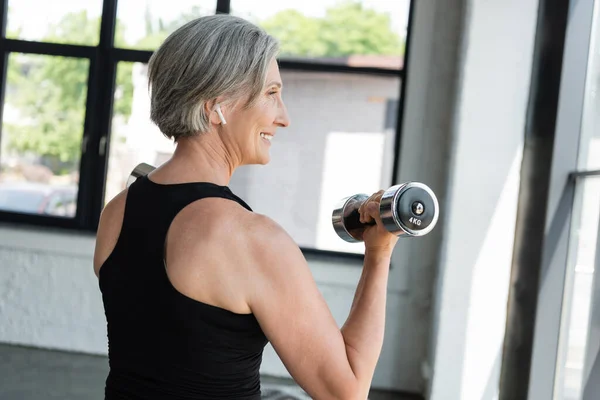 Deportista senior muy alegre en auriculares inalámbricos trabajando con pesas en el gimnasio - foto de stock