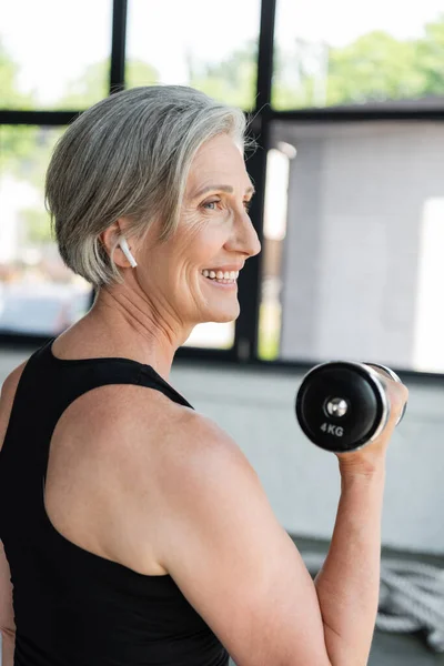 Seitenansicht der überglücklichen Seniorin im drahtlosen Kopfhörer beim Training mit der Hantel im Fitnessstudio — Stockfoto
