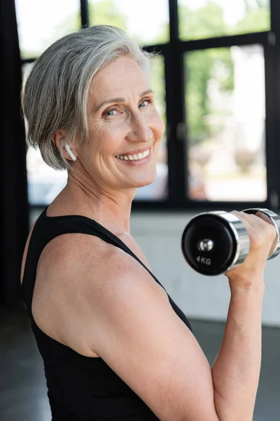 Überglückliche Seniorin mit drahtlosem Kopfhörer trainiert mit Hantel im Fitnessstudio — Stockfoto