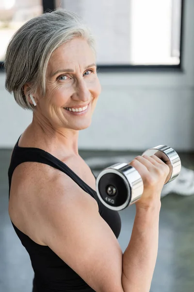 Mujer mayor sonriente en auriculares inalámbricos escuchando música y haciendo ejercicio con mancuerna en el gimnasio - foto de stock