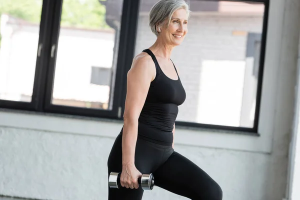 Mujer mayor positiva en auriculares inalámbricos escuchando música mientras hace ejercicio con mancuerna en el gimnasio - foto de stock