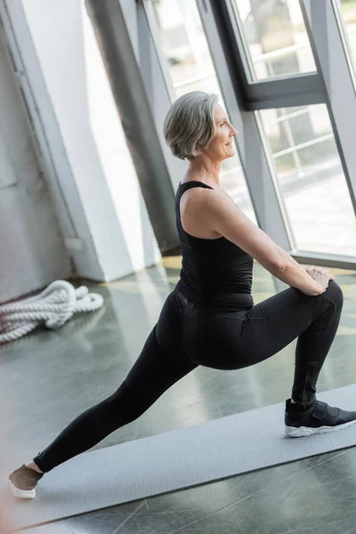 Боковой вид позитивной пожилой женщины в черном платье, делающей упражнения на фитнес-коврике — стоковое фото