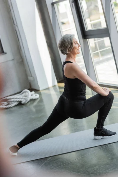 Seitenansicht einer zufriedenen Seniorin in schwarzer Sportbekleidung beim Ausfallschritt auf der Fitnessmatte — Stockfoto