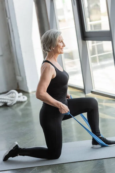 Старшая спортсменка в черных леггинсах и майке, тренирующаяся с полосой сопротивления на коврике для фитнеса — стоковое фото