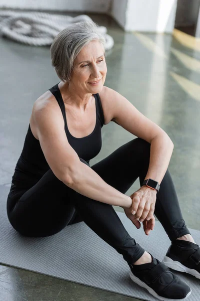 Longitud completa de la mujer mayor feliz en leggings negros y camiseta sin mangas sentado en la colchoneta de fitness - foto de stock