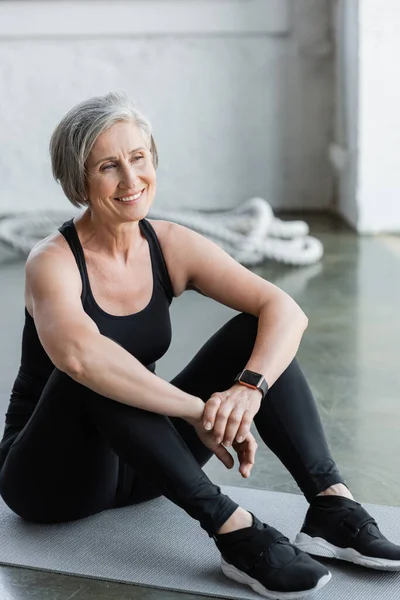 Happy senior woman in black leggings and tank top sitting on fitness mat — Stock Photo