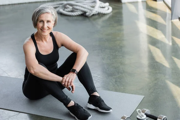 High angle view of happy senior woman in black leggings and tank top sitting on fitness mat — Stock Photo