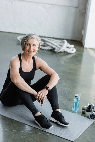 Happy senior woman in black leggings and tank top sitting on fitness mat in gym — Stock Photo