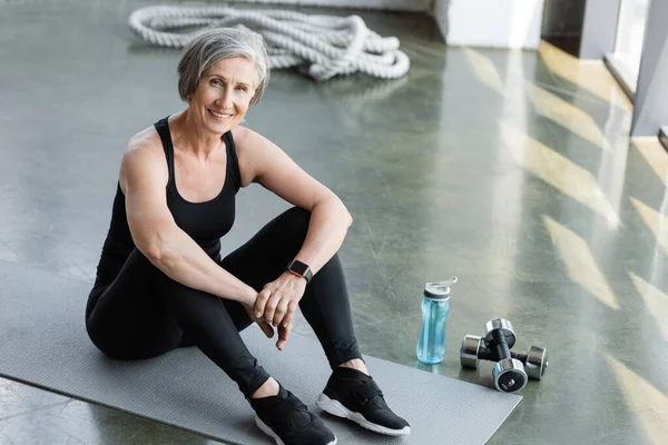 Cheerful senior woman in black leggings and tank top sitting on fitness mat in gym — Stock Photo