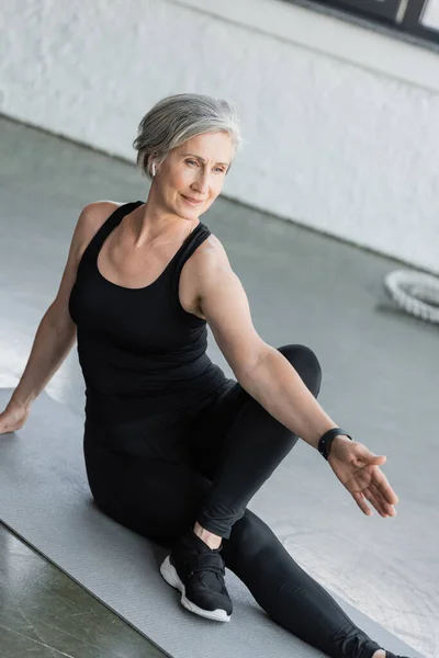 Comprimento total da mulher sênior em leggings pretos e top tanque de exercício no tapete de fitness no ginásio — Fotografia de Stock