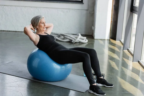 Comprimento total da mulher sênior em leggings pretos e top tanque de exercício na bola de fitness no ginásio — Fotografia de Stock