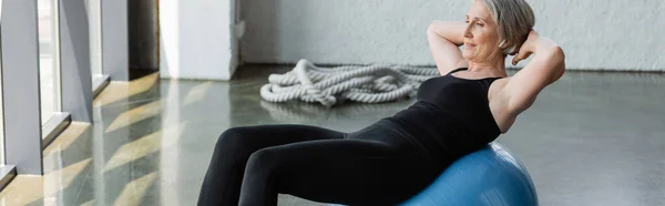 Mulher idosa em leggings pretos e top tanque de exercício na bola de fitness no ginásio, banner — Fotografia de Stock