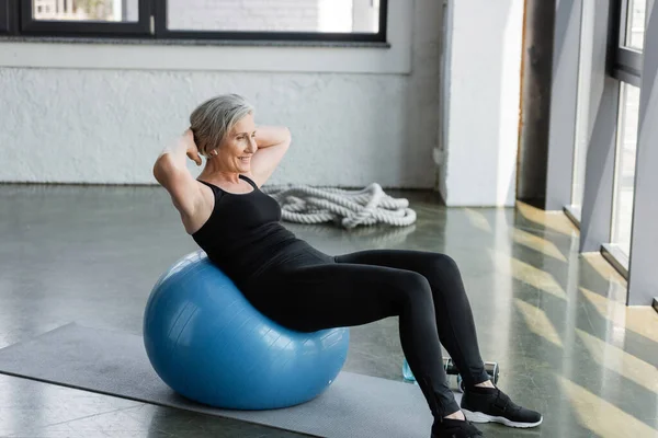 Comprimento total da mulher idosa feliz em leggings pretos e top do tanque exercitando-se na bola de fitness no ginásio — Fotografia de Stock
