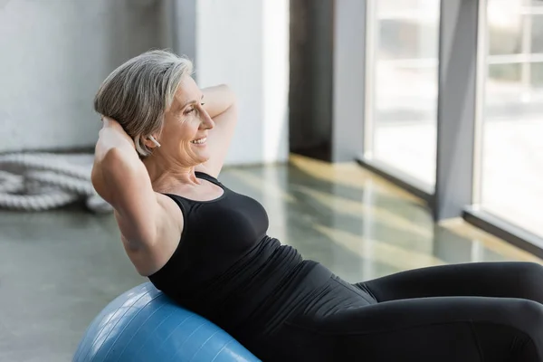 Donna anziana positiva in leggings neri e canotta esercizio su palla fitness blu — Foto stock