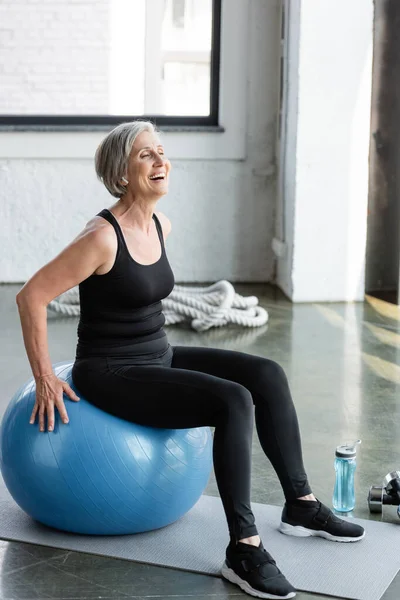 Full length of excited senior woman in black leggings and tank top exercising on blue fitness ball — Stock Photo