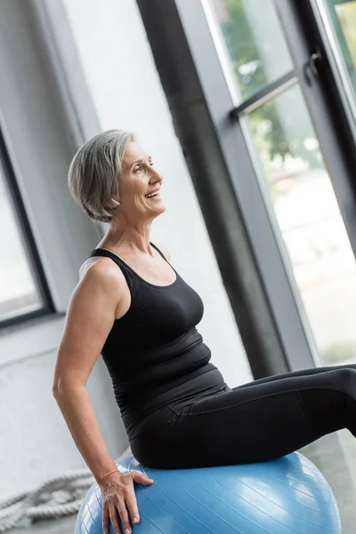 Femme âgée ravie en leggings noirs et débardeur exercice sur boule de fitness bleue — Photo de stock