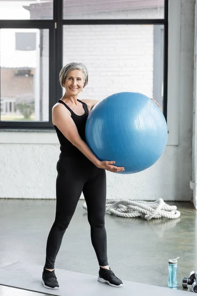 Mujer mayor llena de alegría en leggings negro y camiseta con la celebración de la bola de fitness azul - foto de stock