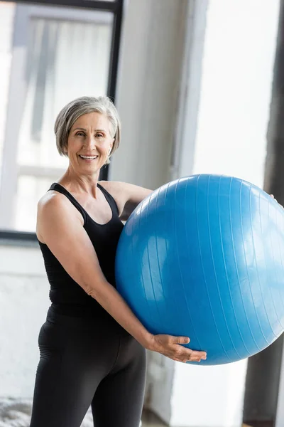 Mujer mayor feliz en ropa deportiva negro sosteniendo pelota de fitness azul mientras hace ejercicio en el gimnasio - foto de stock