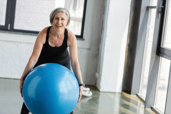 Mujer senior excitada en ropa deportiva negra sosteniendo la pelota de fitness azul mientras hace ejercicio en el gimnasio - foto de stock