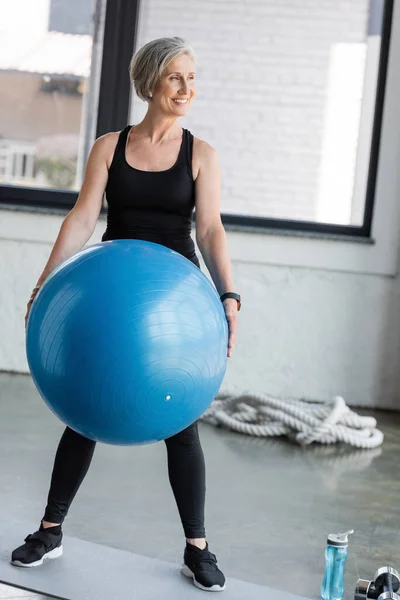 Полная длина веселой пожилой женщины в черной спортивной одежде проведение синий мяч фитнес во время тренировки в тренажерном зале — стоковое фото