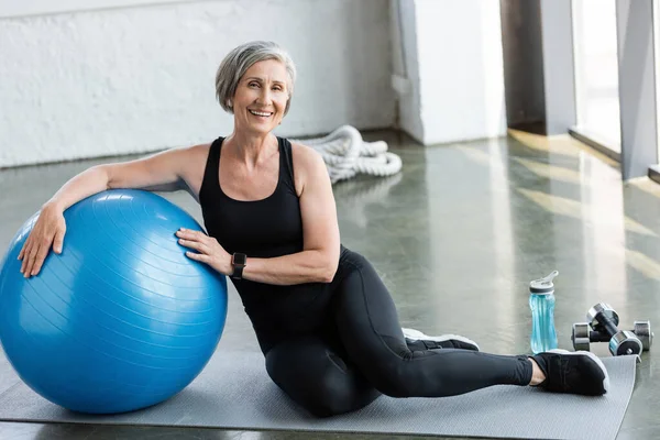 Longitud completa de la mujer mayor feliz en leggings negros y camiseta sin mangas sentado cerca de la pelota de fitness en el gimnasio - foto de stock