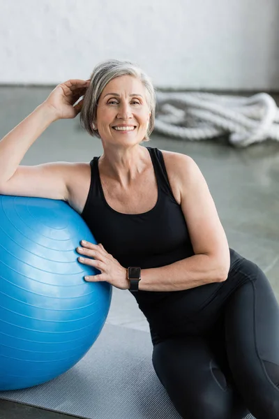 Mujer mayor feliz en leggings negros y camiseta sin mangas sentado cerca de la pelota de fitness azul en el gimnasio - foto de stock