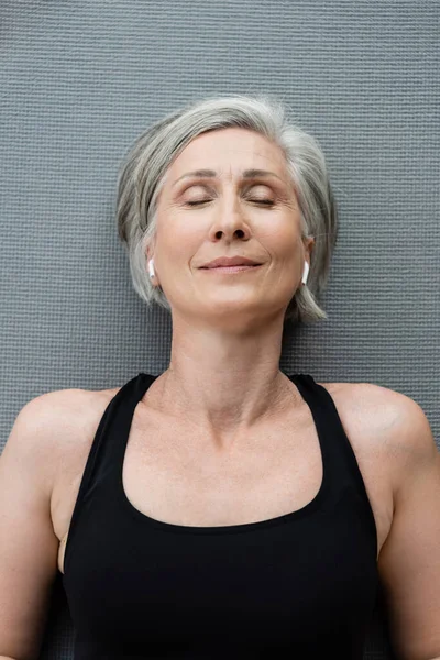 Vista superior de la mujer mayor complacida en los auriculares inalámbricos que yacen en la estera de fitness - foto de stock