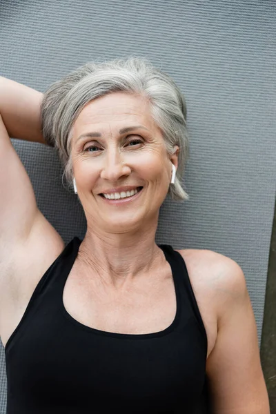 Draufsicht auf glückliche Seniorin in drahtlosen Kopfhörern, die auf Fitnessmatte liegt — Stockfoto