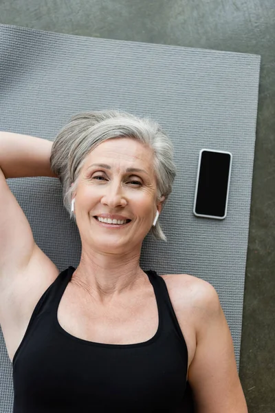 Vista superior da mulher sênior satisfeito em fones de ouvido sem fio deitado perto do smartphone com tela em branco no tapete de fitness — Fotografia de Stock
