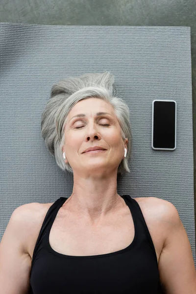 Vista superior da mulher idosa feliz em fones de ouvido sem fio que jazem perto do smartphone com tela em branco no tapete de fitness — Fotografia de Stock
