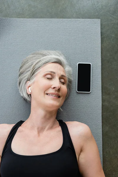 Vista superior de la mujer mayor sonriente en los auriculares inalámbricos que se encuentran cerca del teléfono inteligente con pantalla en blanco en la estera de fitness - foto de stock