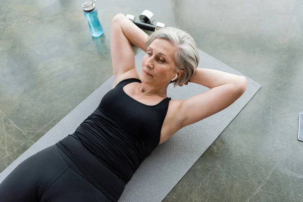 Vista de ángulo alto de la mujer mayor en auriculares inalámbricos haciendo abdominales ejercicio en la estera de fitness - foto de stock