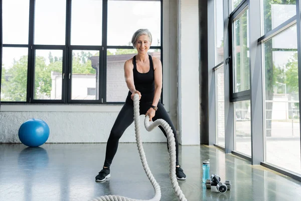 Longitud completa de la mujer mayor feliz que trabaja con cuerdas de batalla cerca de pesas en el gimnasio - foto de stock
