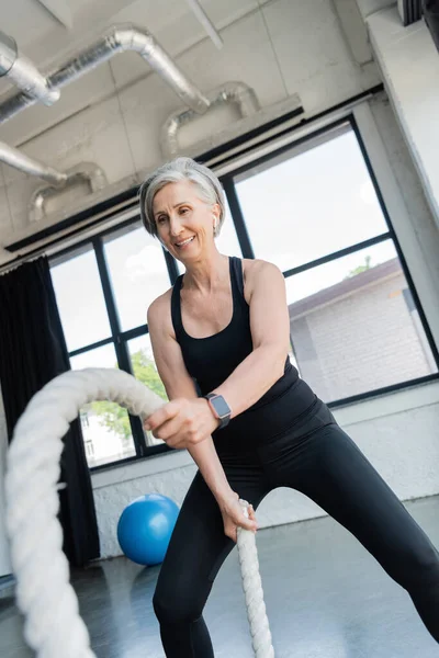 Happy senior sportswoman with grey hair working out with battle ropes in gym — Stock Photo