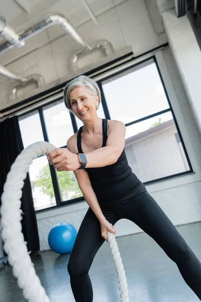 Cheerful senior sportswoman with grey hair working out with battle ropes in gym — Stock Photo